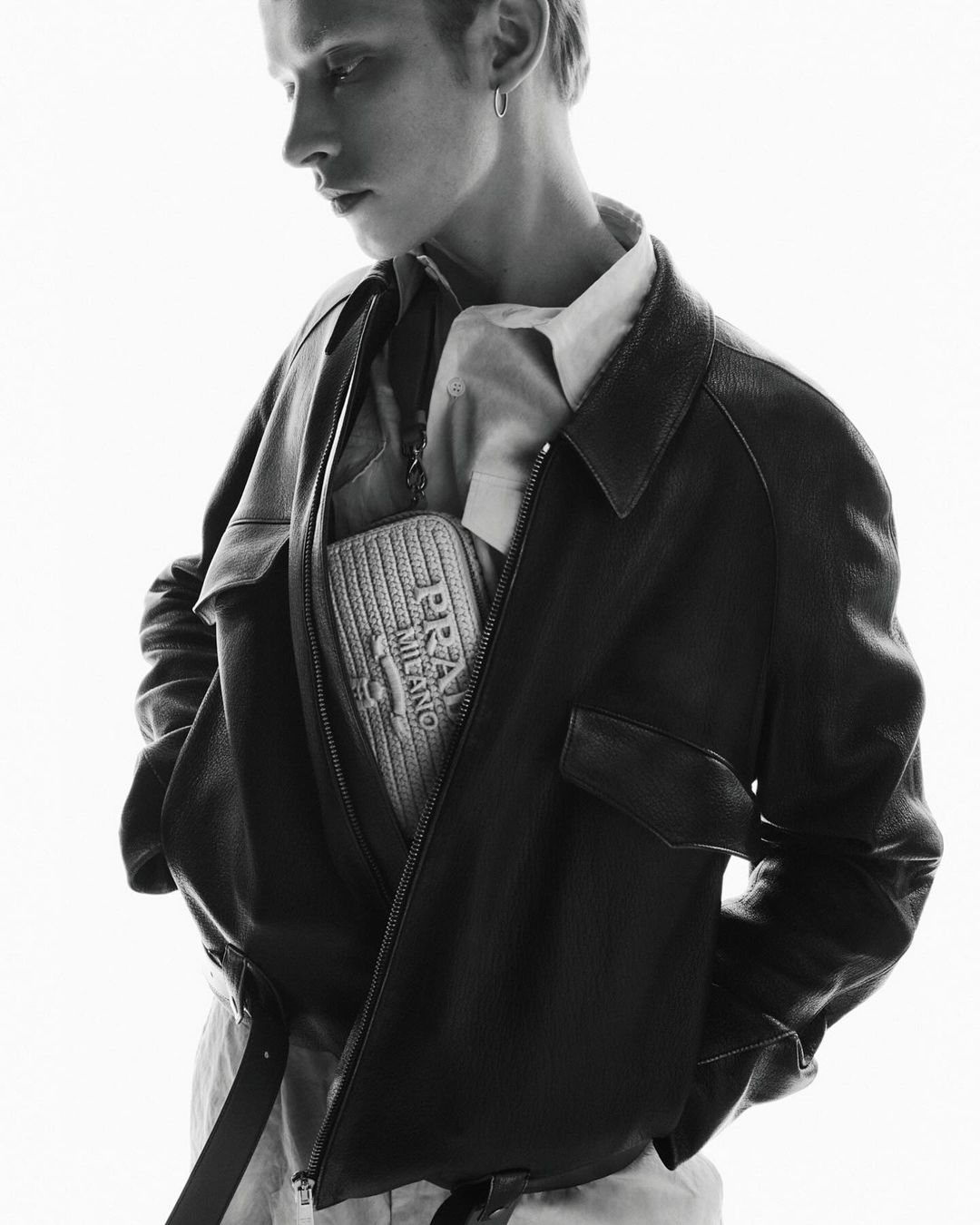 Monochrome fashion portrait of a young adult male wearing a dark bomber jacket with a 'Prada Milano' bag in an indoor setting.