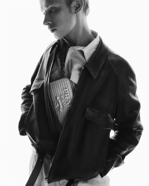 Monochrome fashion portrait of a young adult male wearing a dark bomber jacket with a 'Prada Milano' bag in an indoor setting.