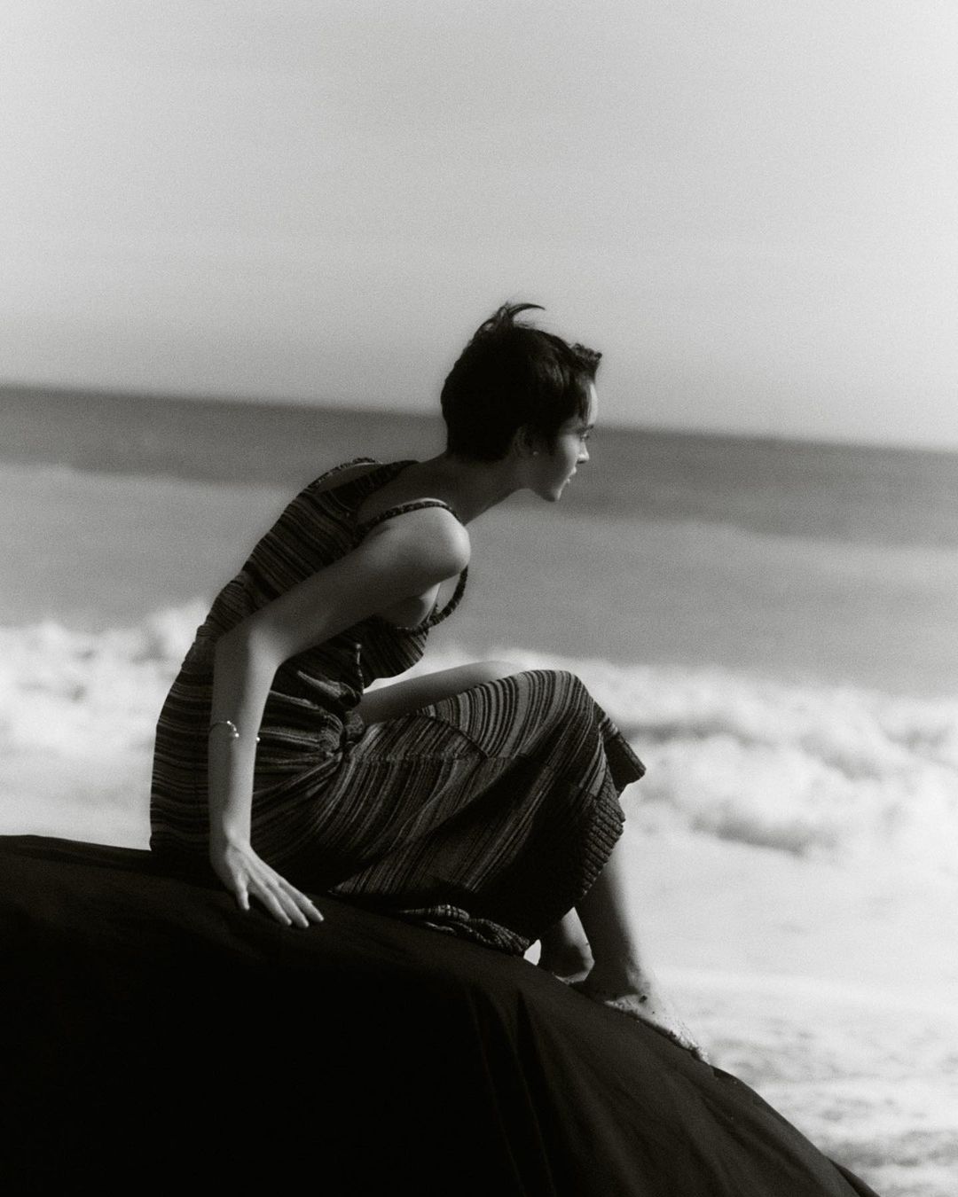 Monochrome photograph of a woman in a dress sitting on a rock at the beach looking out at the water, bathed in natural light.