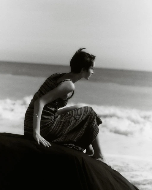 Monochrome photograph of a woman in a dress sitting on a rock at the beach looking out at the water, bathed in natural light.