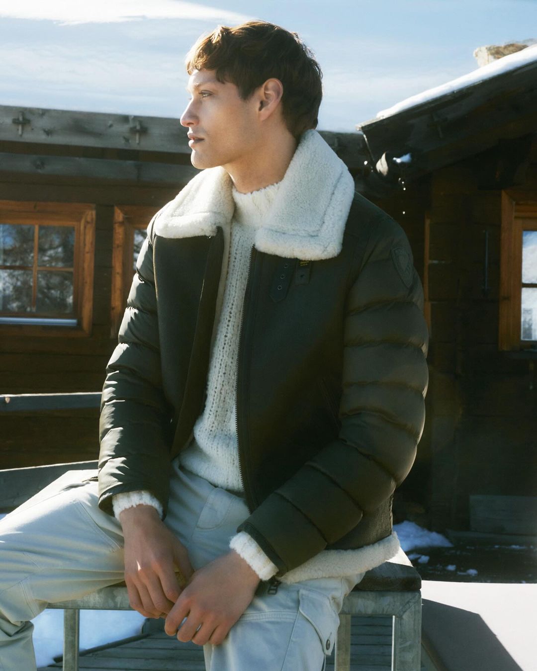 A man in a shearling-trimmed puffer jacket looks to the left against a backdrop of a log cabin and snowy environment in an eye-level outdoor shot.