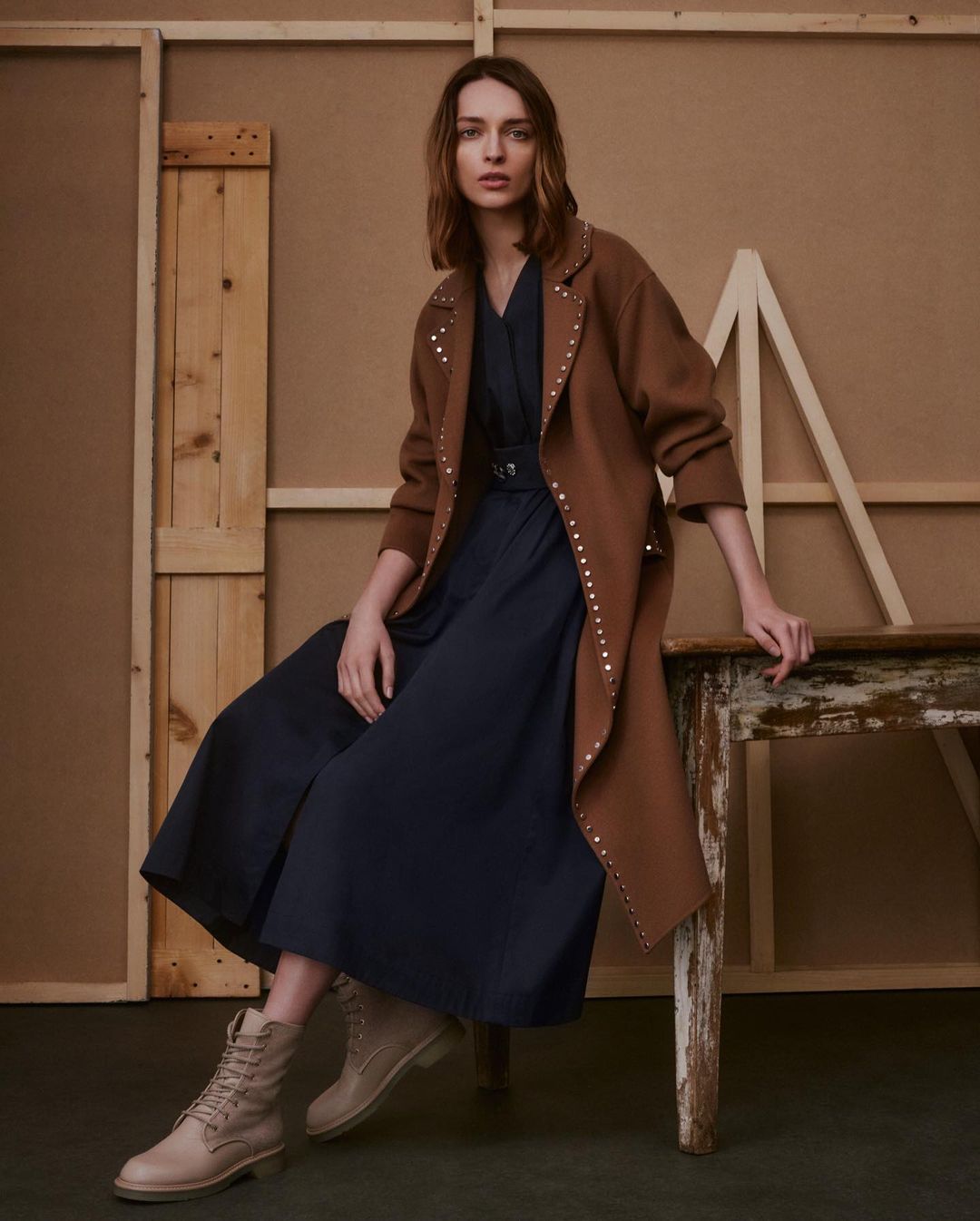 Adult woman in brown studded coat and navy jumpsuit sits on wooden table against tan studio backdrop in fashion photography setting.