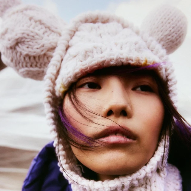 Close-up portrait of a young adult female with dyed purple hair, wearing a knitted bear-ear hat and jacket against a light outdoor backdrop.