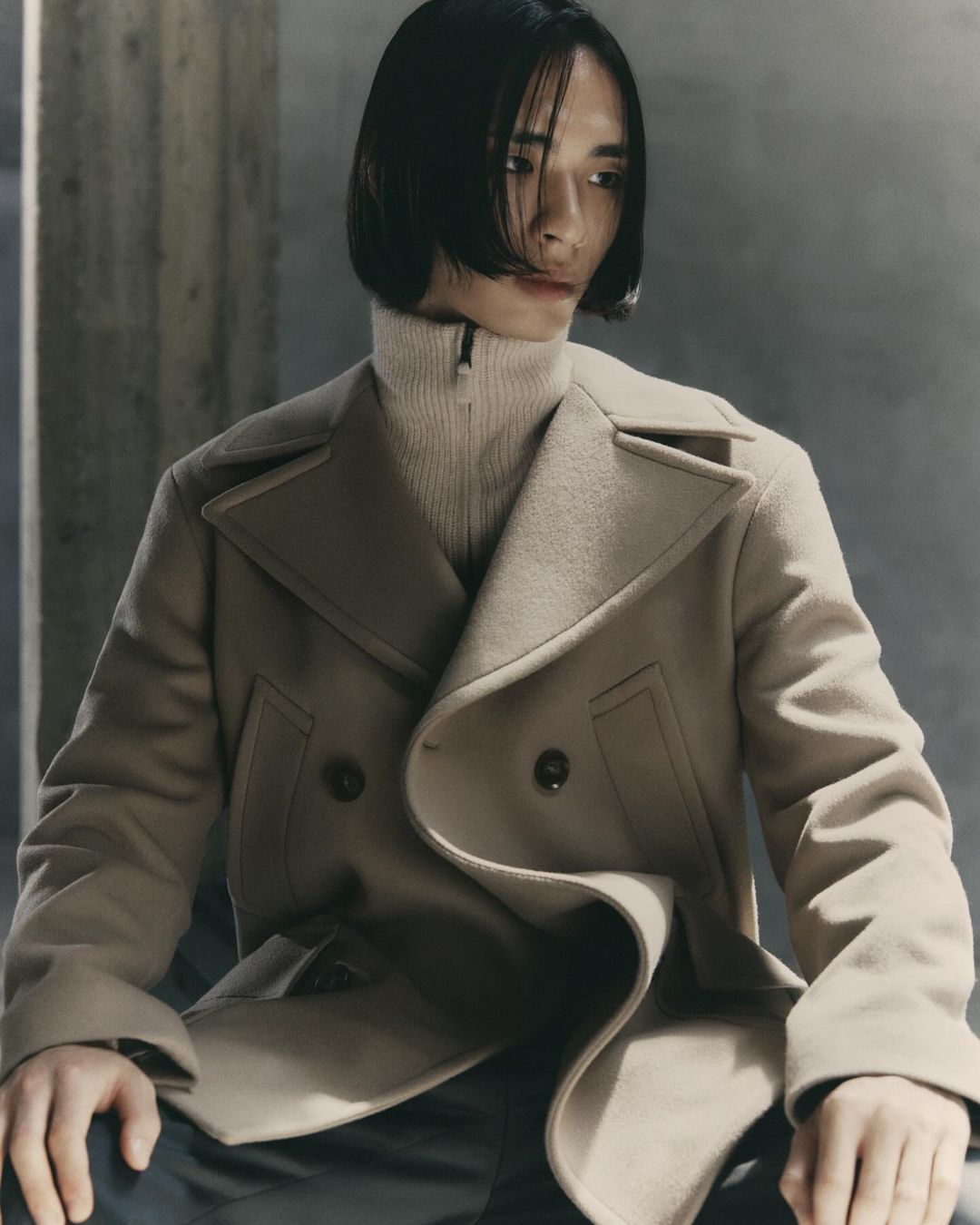 An adult male with dark hair poses indoors wearing a tan double-breasted pea coat over a cream-colored turtleneck sweater, fashion portrait.