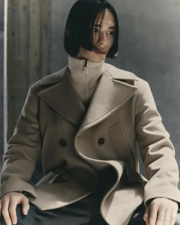 An adult male with dark hair poses indoors wearing a tan double-breasted pea coat over a cream-colored turtleneck sweater, fashion portrait.