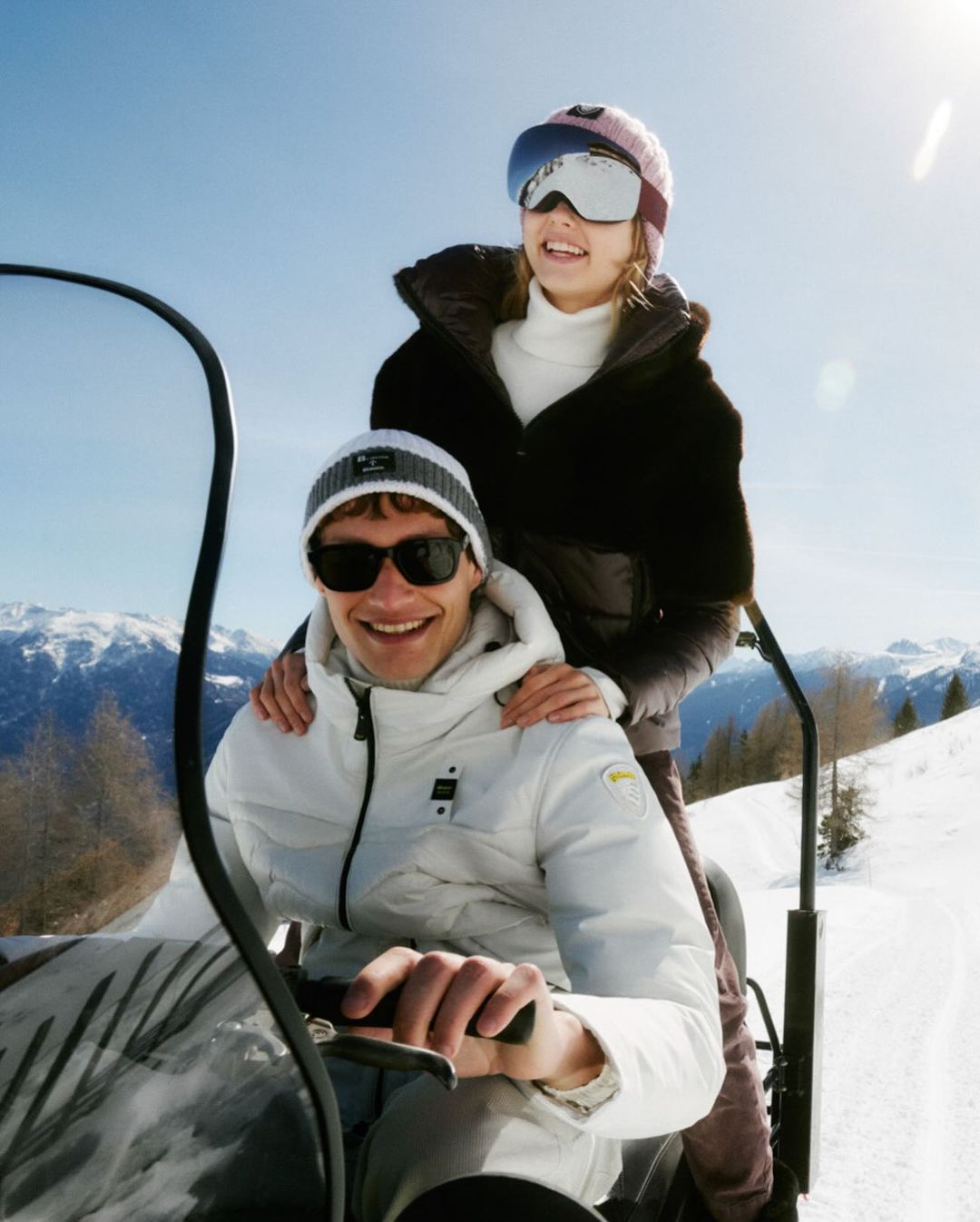 A man and woman on a snowmobile in a snowy mountain landscape with clear skies, wearing winter clothing and goggles, capturing a winter sports scene.