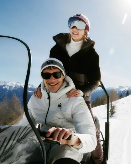 A man and woman on a snowmobile in a snowy mountain landscape with clear skies, wearing winter clothing and goggles, capturing a winter sports scene.