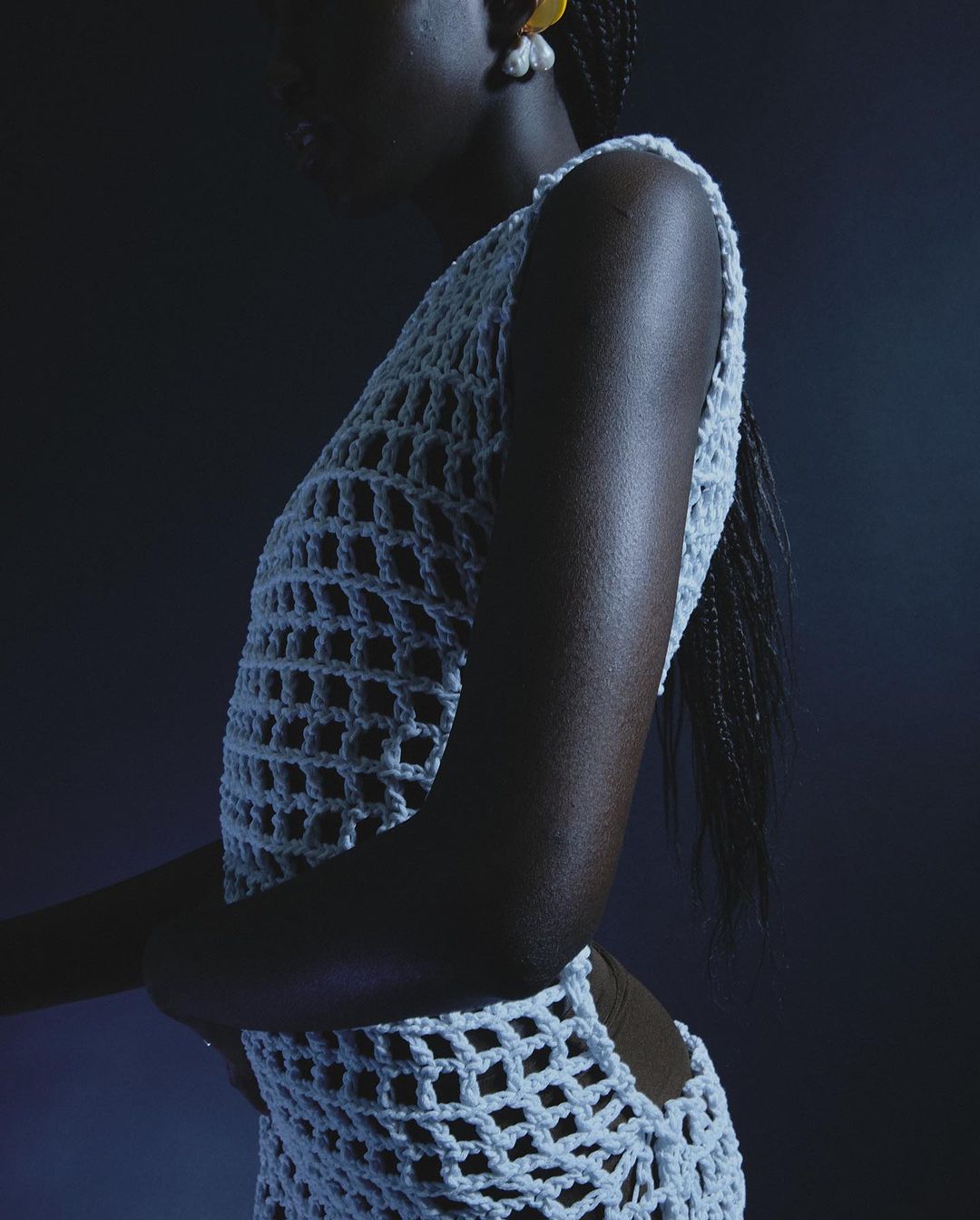 An adult female with dark skin poses in a crochet style garment. She is indoors with a dark background and wears large earrings.