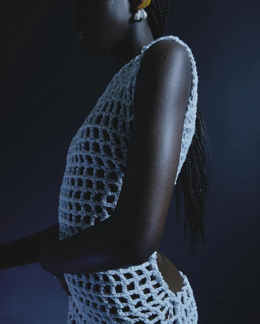 An adult female with dark skin poses in a crochet style garment. She is indoors with a dark background and wears large earrings.