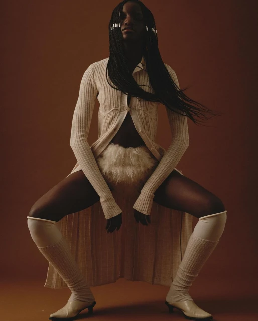 Elegant studio shot of a Black woman posing in a light-colored knit outfit with long braids, against a warm brown backdrop.
