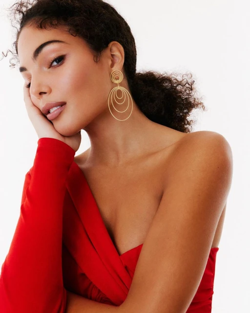Portrait of a dark-haired woman with golden earrings wearing a red, one-shoulder dress against a white background.