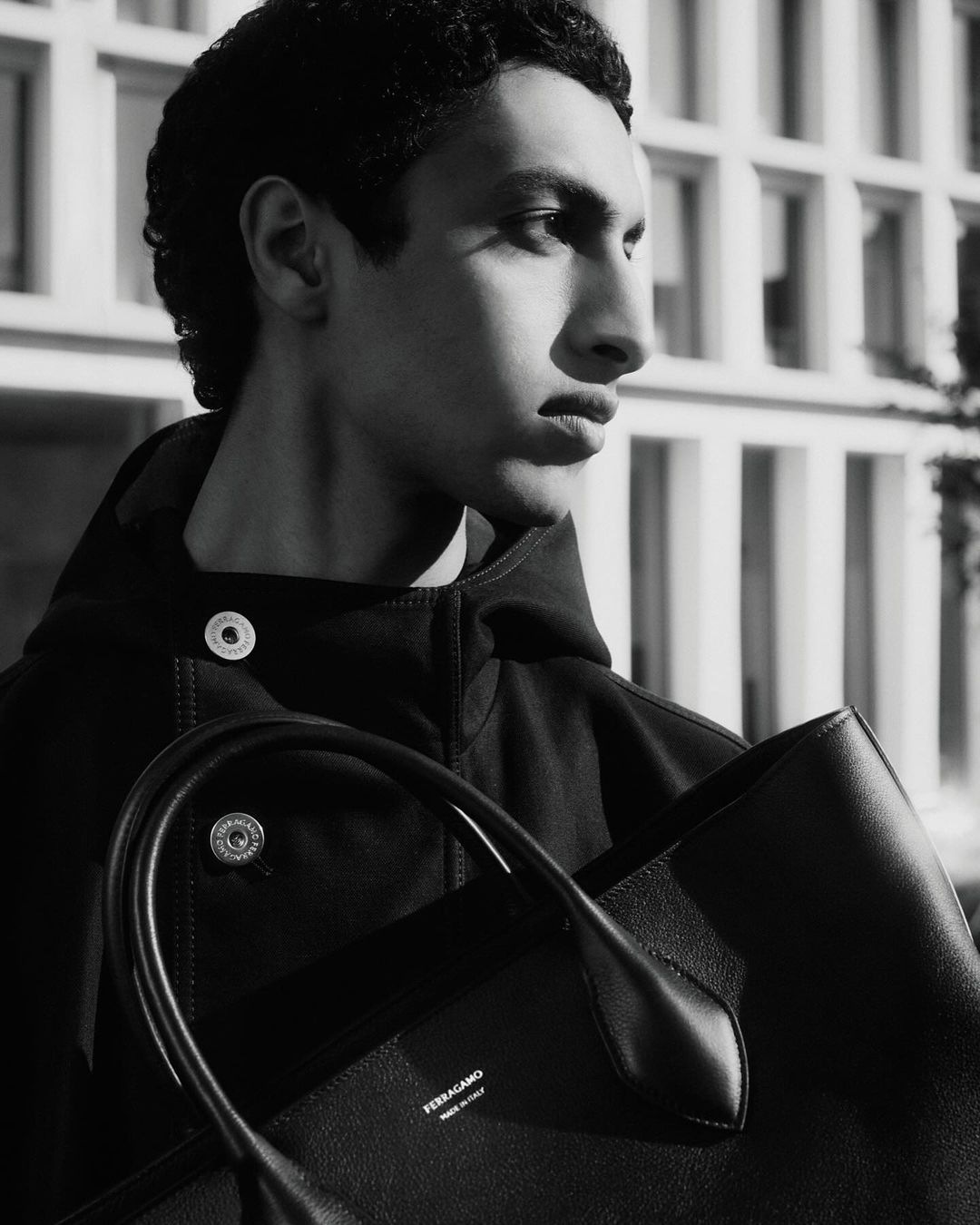 Black-and-white portrait of a young adult male model outdoors, wearing a dark hooded jacket and holding a black leather tote bag.