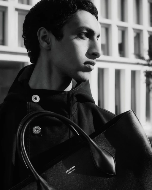 Black-and-white portrait of a young adult male model outdoors, wearing a dark hooded jacket and holding a black leather tote bag.