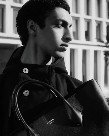 Black-and-white portrait of a young adult male model outdoors, wearing a dark hooded jacket and holding a black leather tote bag.