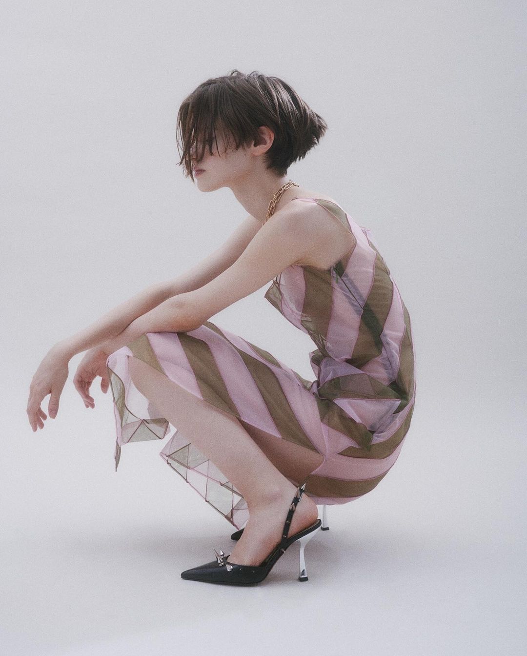 Candid shot of a person with short hair, posing in a striped dress and black heels against a plain background in a studio setting.