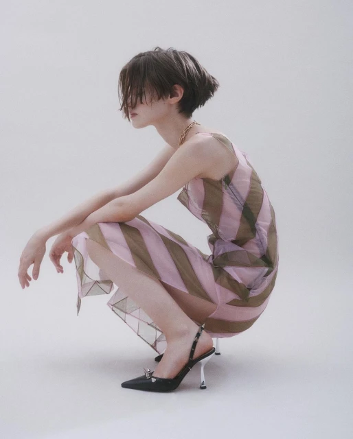 Candid shot of a person with short hair, posing in a striped dress and black heels against a plain background in a studio setting.