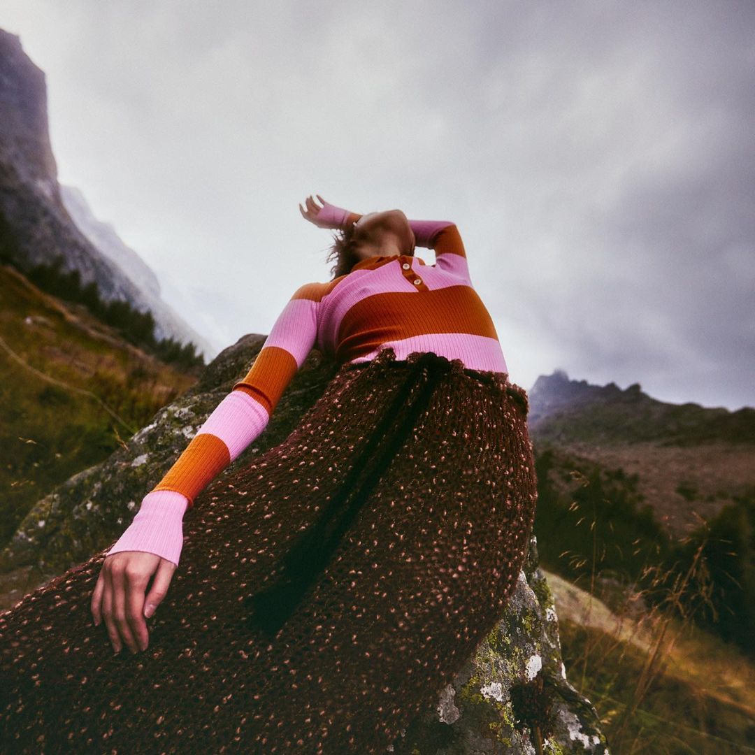 Woman in brown pleated skirt and pink striped top posing on rock in mountainous landscape under an overcast sky. Fashion shot.