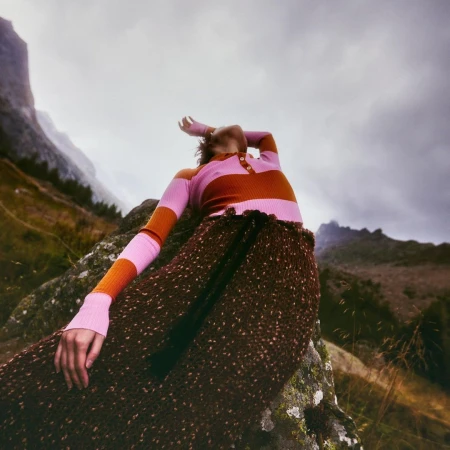 Woman in brown pleated skirt and pink striped top posing on rock in mountainous landscape under an overcast sky. Fashion shot.