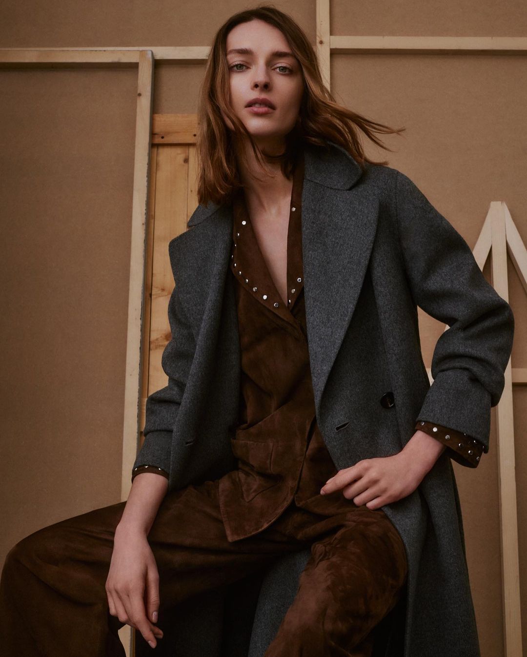 A stylish adult female is captured indoors, seated and posing in a gray coat over a brown suede-like outfit, against a muted background with artistic panels.