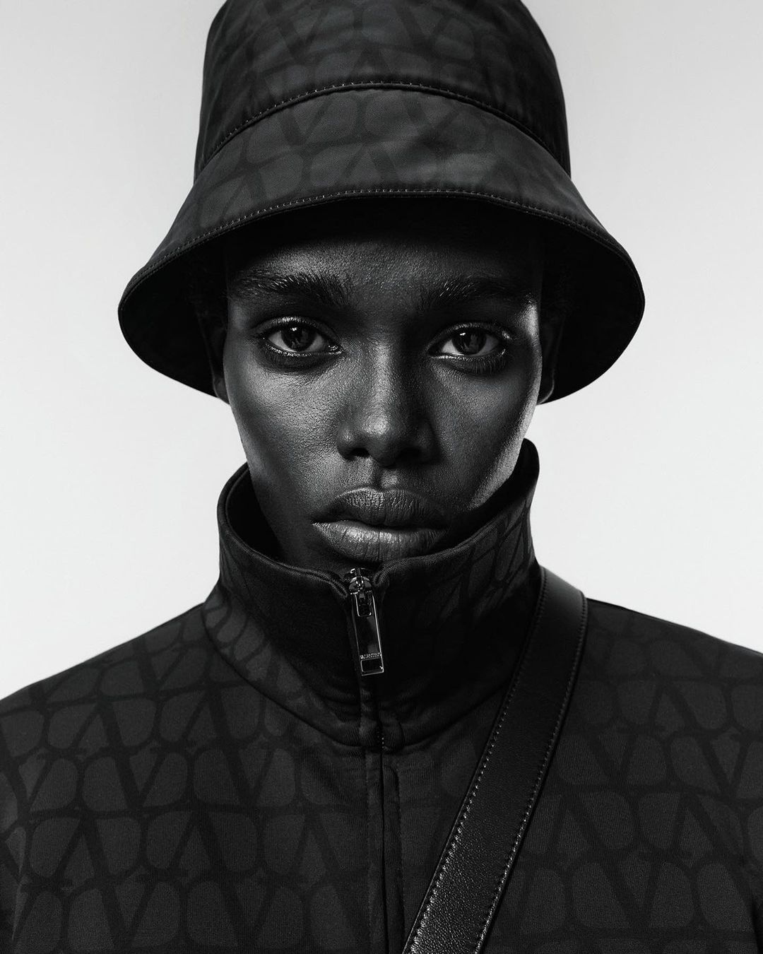 Portrait of a woman with dark skin wearing a black patterned bucket hat and jacket, a shoulder strap, posing against a neutral background.