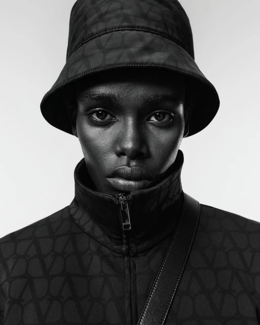 Portrait of a woman with dark skin wearing a black patterned bucket hat and jacket, a shoulder strap, posing against a neutral background.