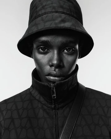 Portrait of a woman with dark skin wearing a black patterned bucket hat and jacket, a shoulder strap, posing against a neutral background.