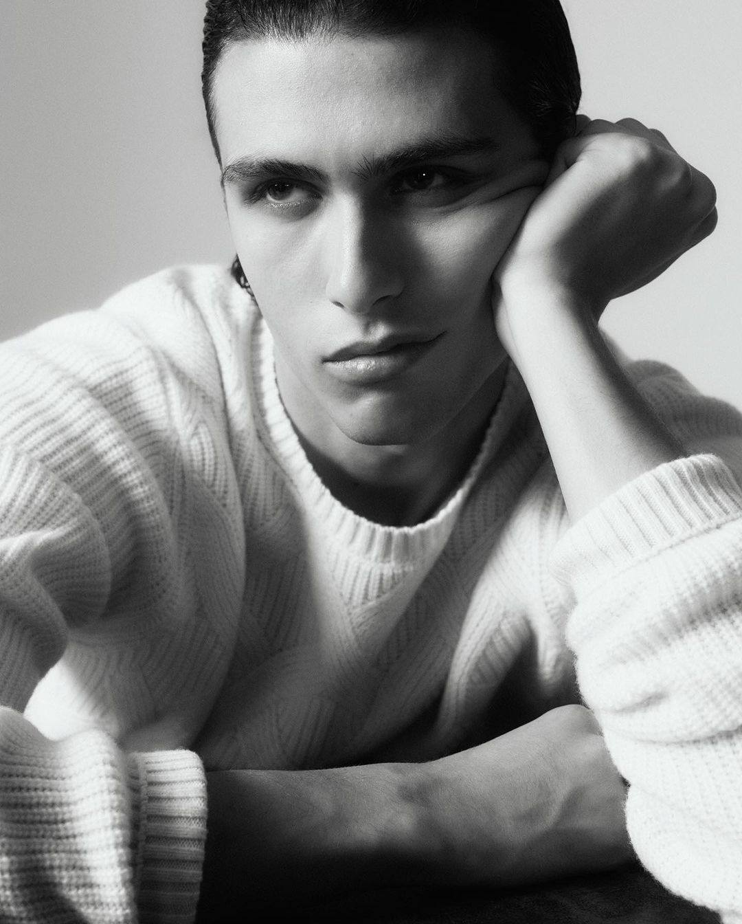 Portrait of an adult male with dark hair wearing a white sweater, resting his head on his hand in a light grey indoor setting.
