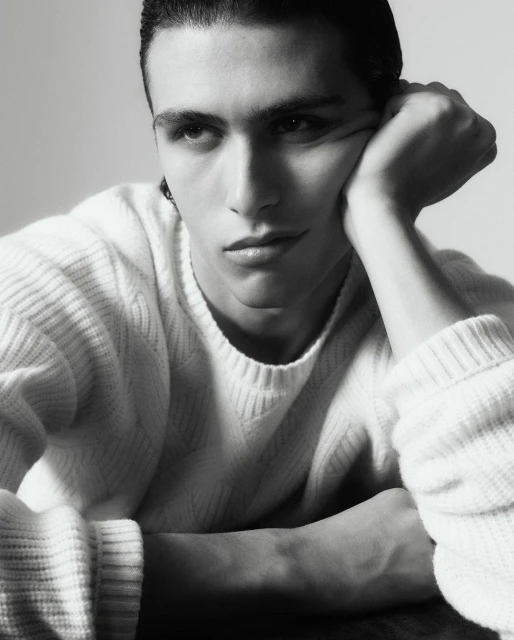 Portrait of an adult male with dark hair wearing a white sweater, resting his head on his hand in a light grey indoor setting.