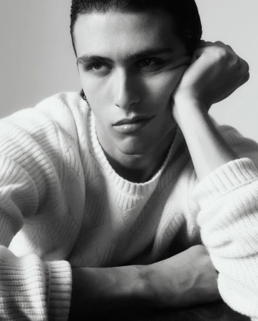 Portrait of an adult male with dark hair wearing a white sweater, resting his head on his hand in a light grey indoor setting.