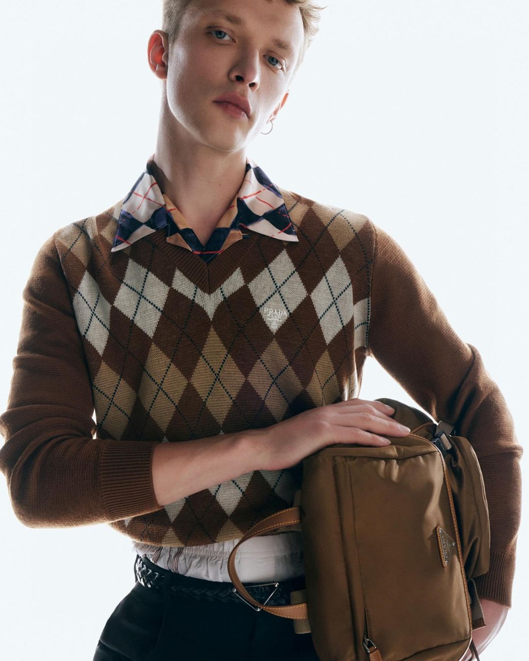 Portrait of a fair-skinned young adult male in a brown argyle sweater holding a backpack in a brightly lit indoor setting.