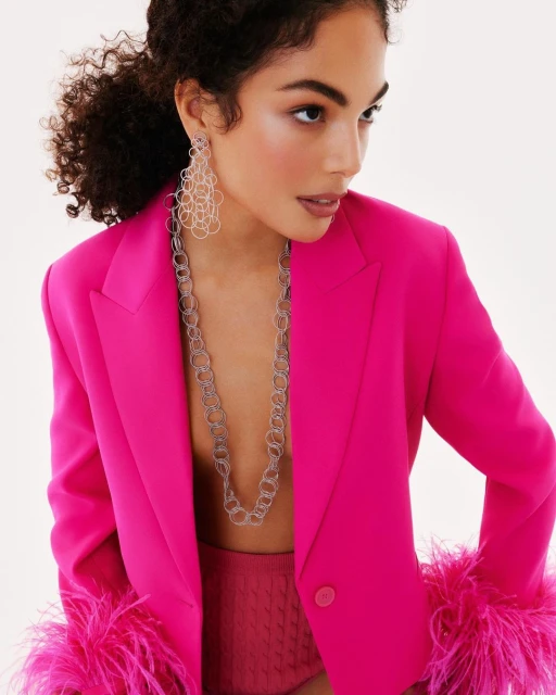 Portrait of a woman in a hot pink suit with feathered sleeves, silver statement jewelry, and a clean, bright background.