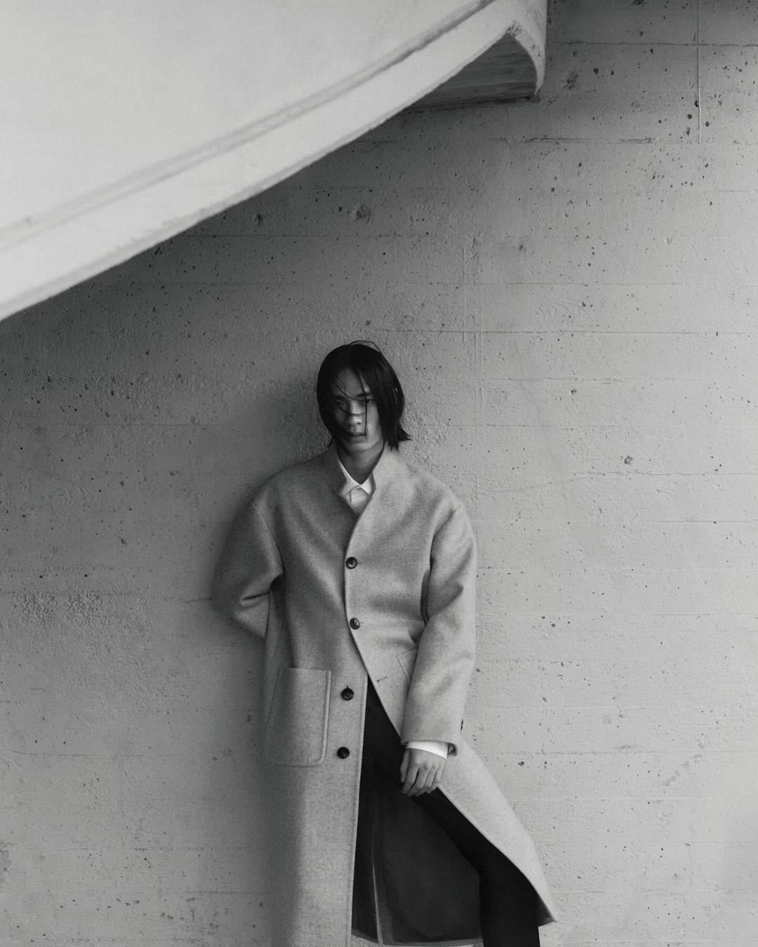 Monochrome fashion shot of a long-haired person in a neutral coat posing indoors against a textured grey wall with angled architectural features.
