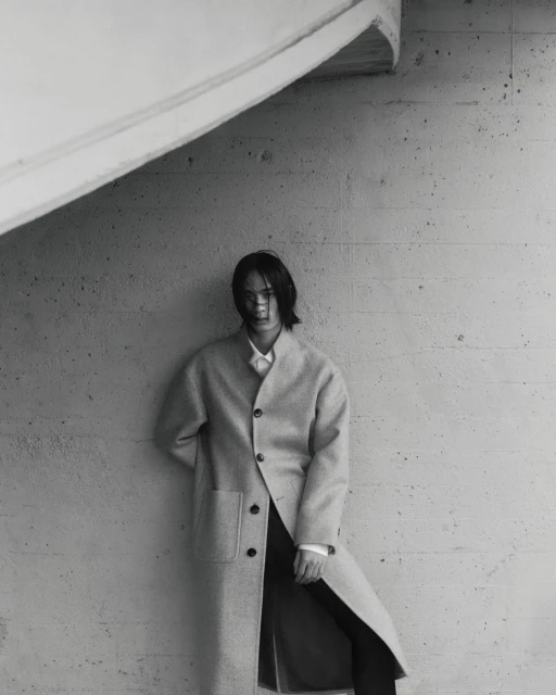 Monochrome fashion shot of a long-haired person in a neutral coat posing indoors against a textured grey wall with angled architectural features.