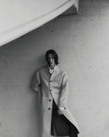 Monochrome fashion shot of a long-haired person in a neutral coat posing indoors against a textured grey wall with angled architectural features.