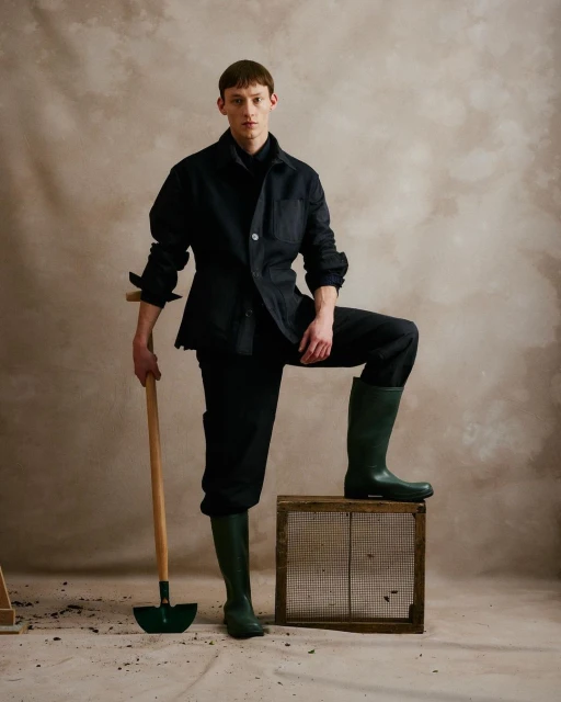 A man poses with a shovel, wearing dark clothing and green rubber boots, standing next to a wooden crate in a studio setting.