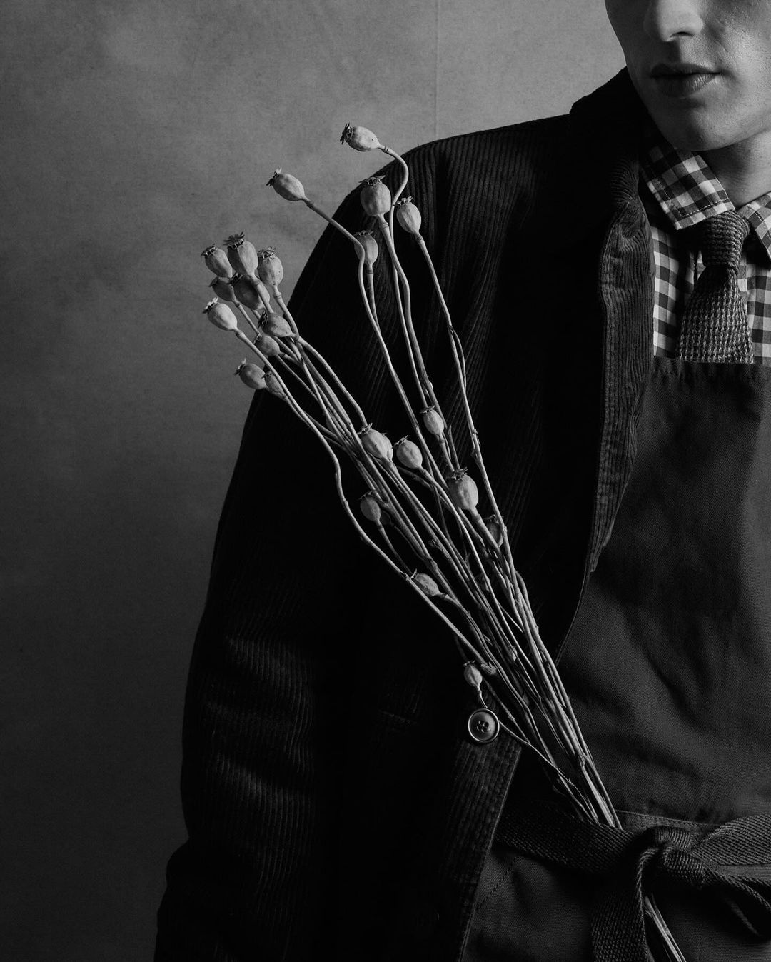 Monochrome portrait of a young man holding dried poppy seed heads, wearing a dark corduroy jacket and checkered shirt in a studio setting.