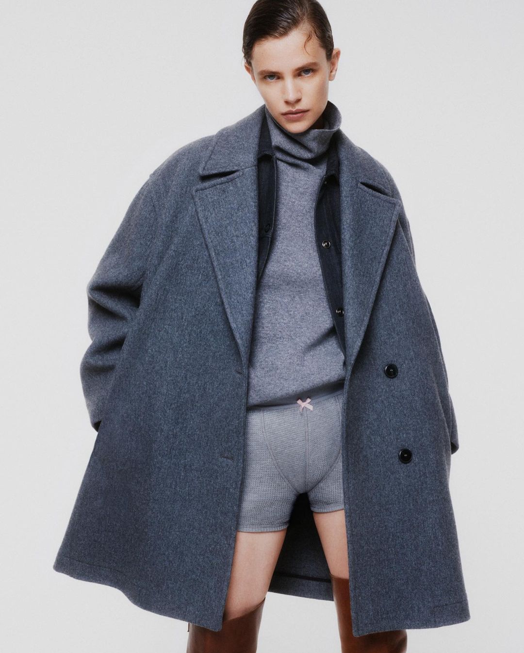 A woman with neutral expression stands in a grey coat and shorts with brown boots against a white backdrop in a fashion studio shot.