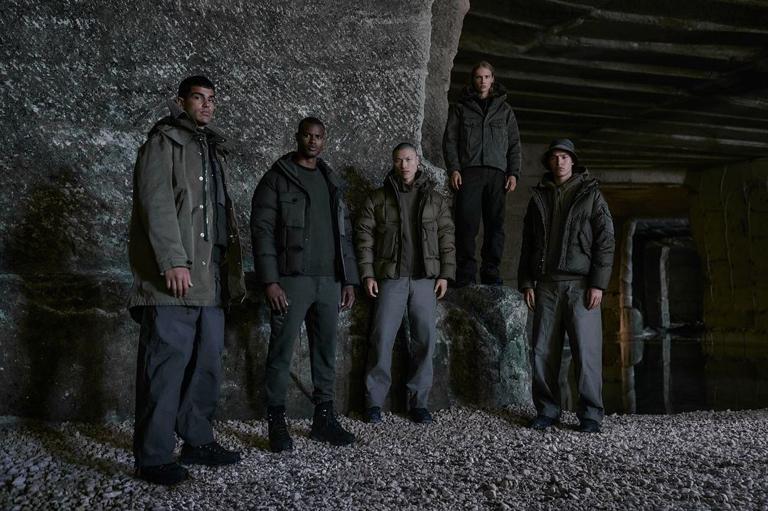 Group of five diverse adult models in olive green jackets and pants posing against a rocky wall in a dimly lit, underground setting.