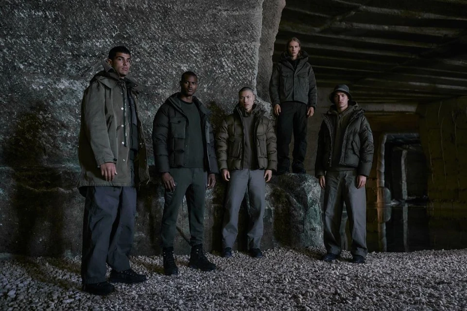 Group of five diverse adult models in olive green jackets and pants posing against a rocky wall in a dimly lit, underground setting.
