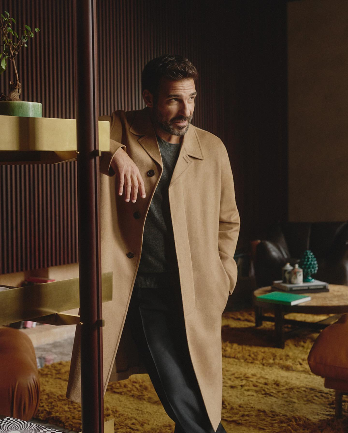 Adult man in tan trench coat poses indoors near mid-century modern shelving and furniture in a warmly lit, stylish interior setting.