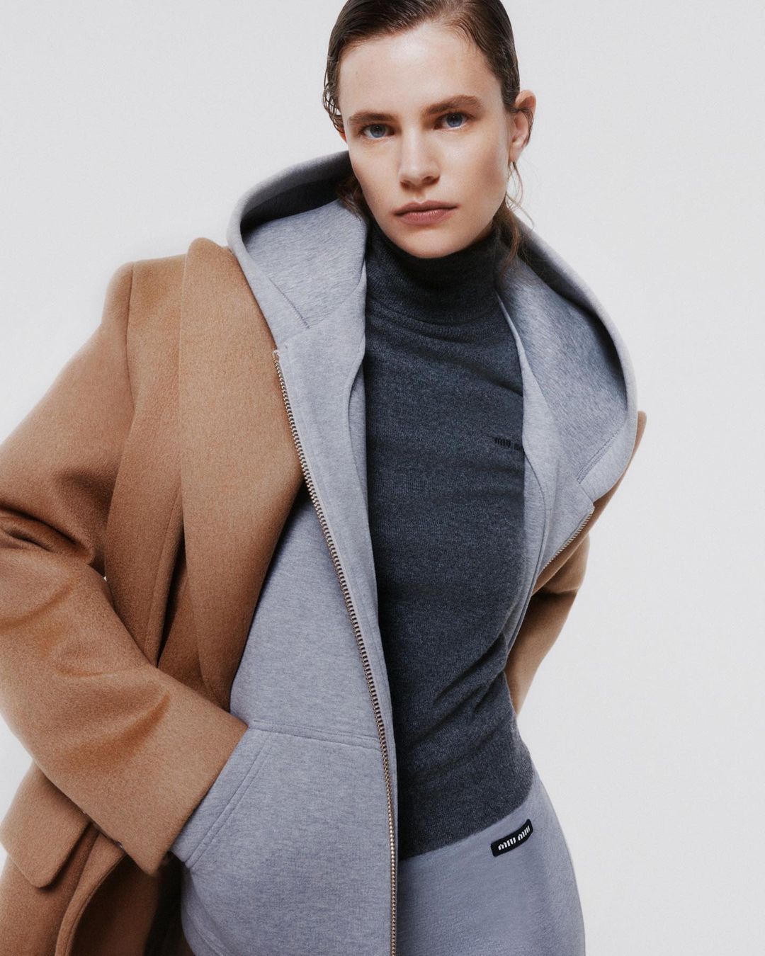 Woman poses indoors wearing a beige coat over a gray hooded sweatshirt and charcoal turtleneck, with a neutral backdrop.