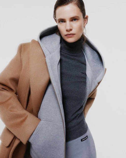 Woman poses indoors wearing a beige coat over a gray hooded sweatshirt and charcoal turtleneck, with a neutral backdrop.