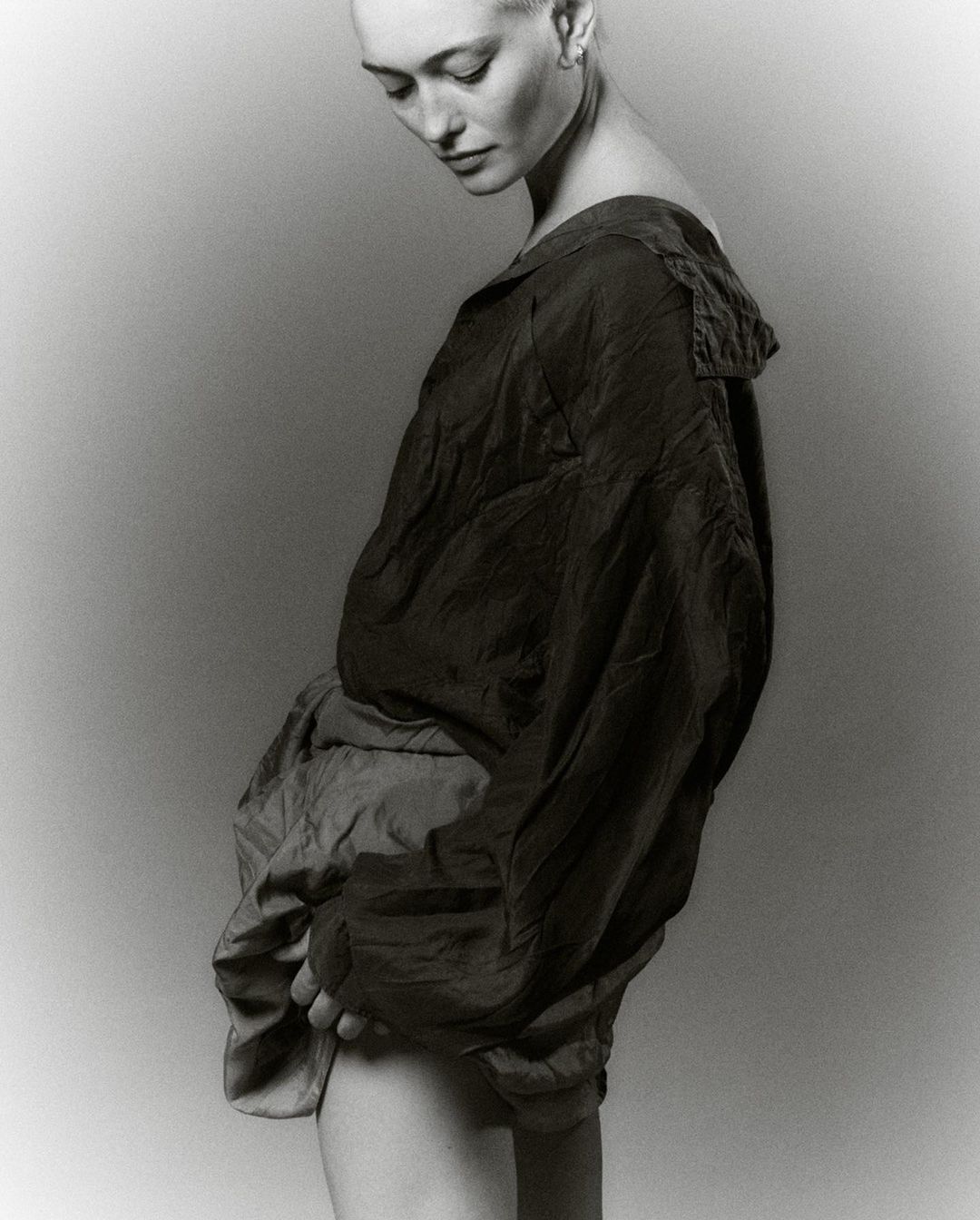 Monochrome portrait of a bald woman in a draped, collared top and shorts, gazing downwards in a studio setting with soft, neutral lighting.