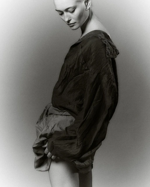 Monochrome portrait of a bald woman in a draped, collared top and shorts, gazing downwards in a studio setting with soft, neutral lighting.