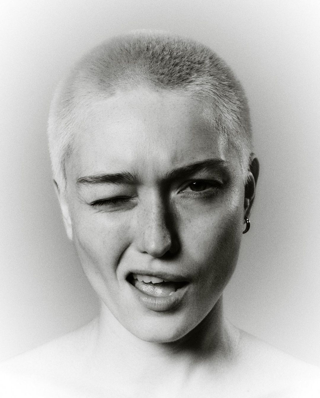 Close-up portrait of a person with a shaved head winking and smiling against a vignette background by Gautier Pellegrin
