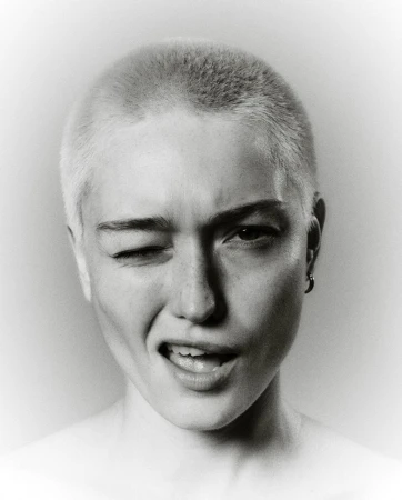 Close-up portrait of a person with a shaved head winking and smiling against a vignette background by Gautier Pellegrin