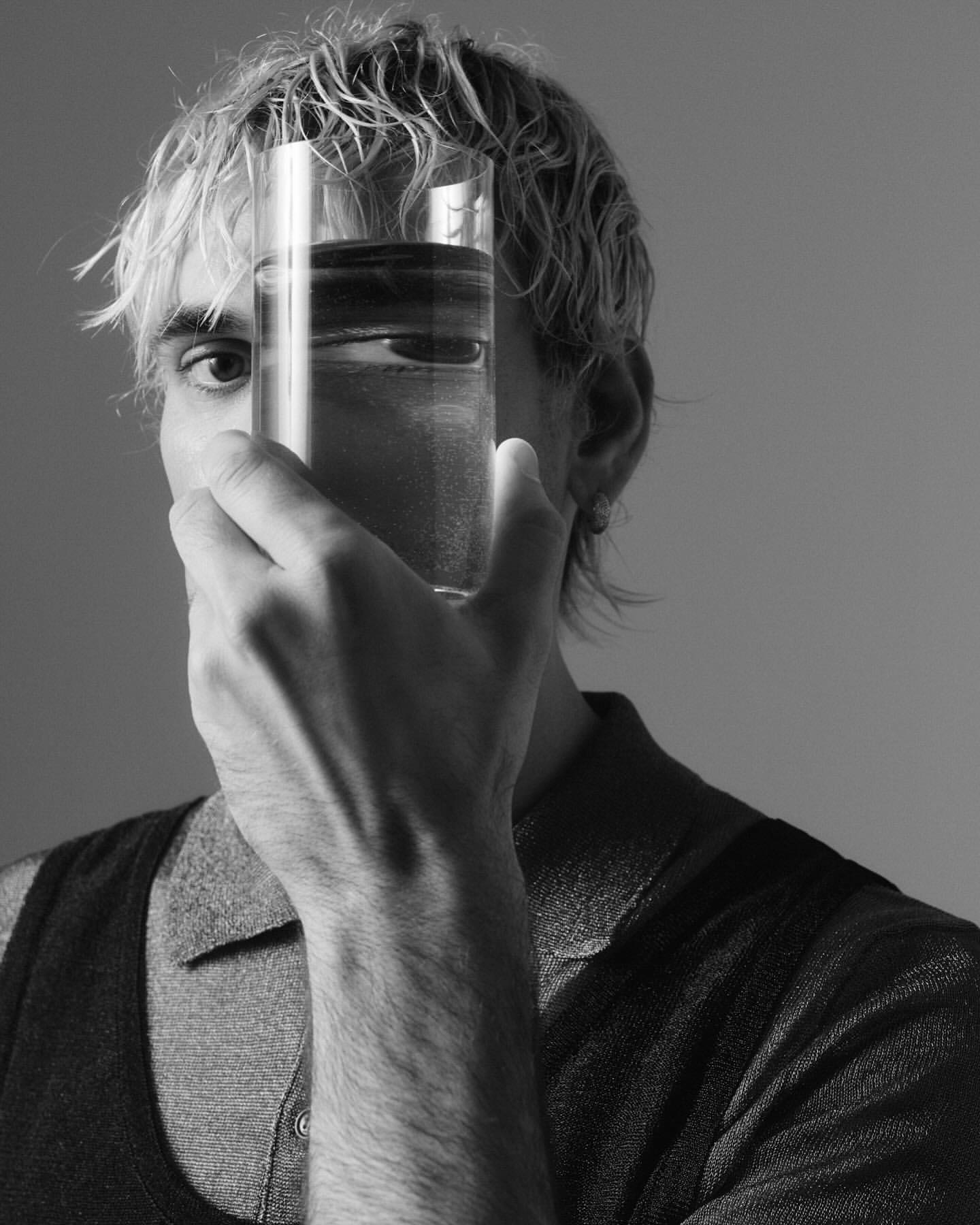 Monochromatic portrait of a blonde-haired man holding a glass of water that distorts his face, set against a neutral background.