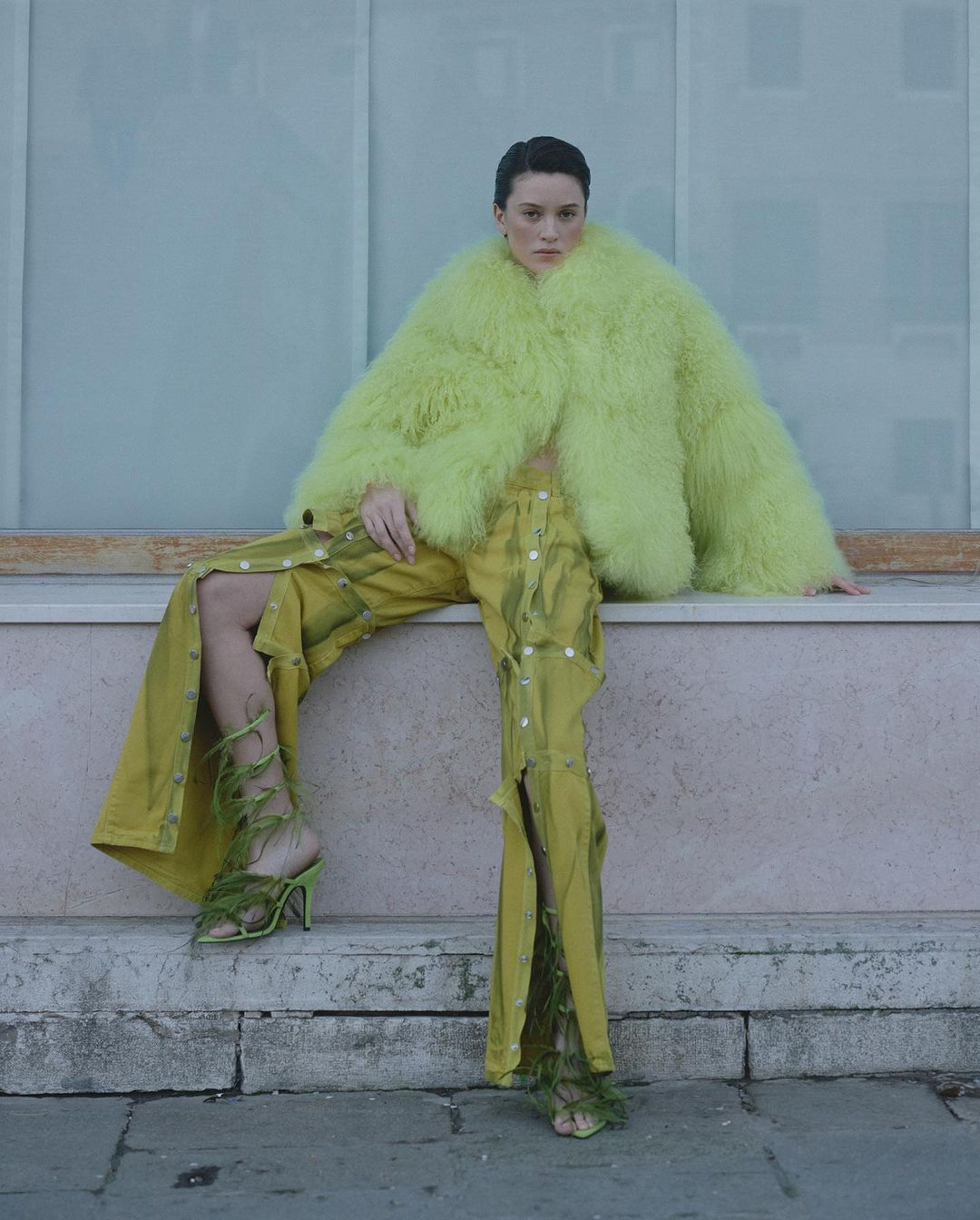 Woman in vibrant faux fur coat and avant-garde olive pants poses outdoors, completed with lace-up heels, against a neutral backdrop.