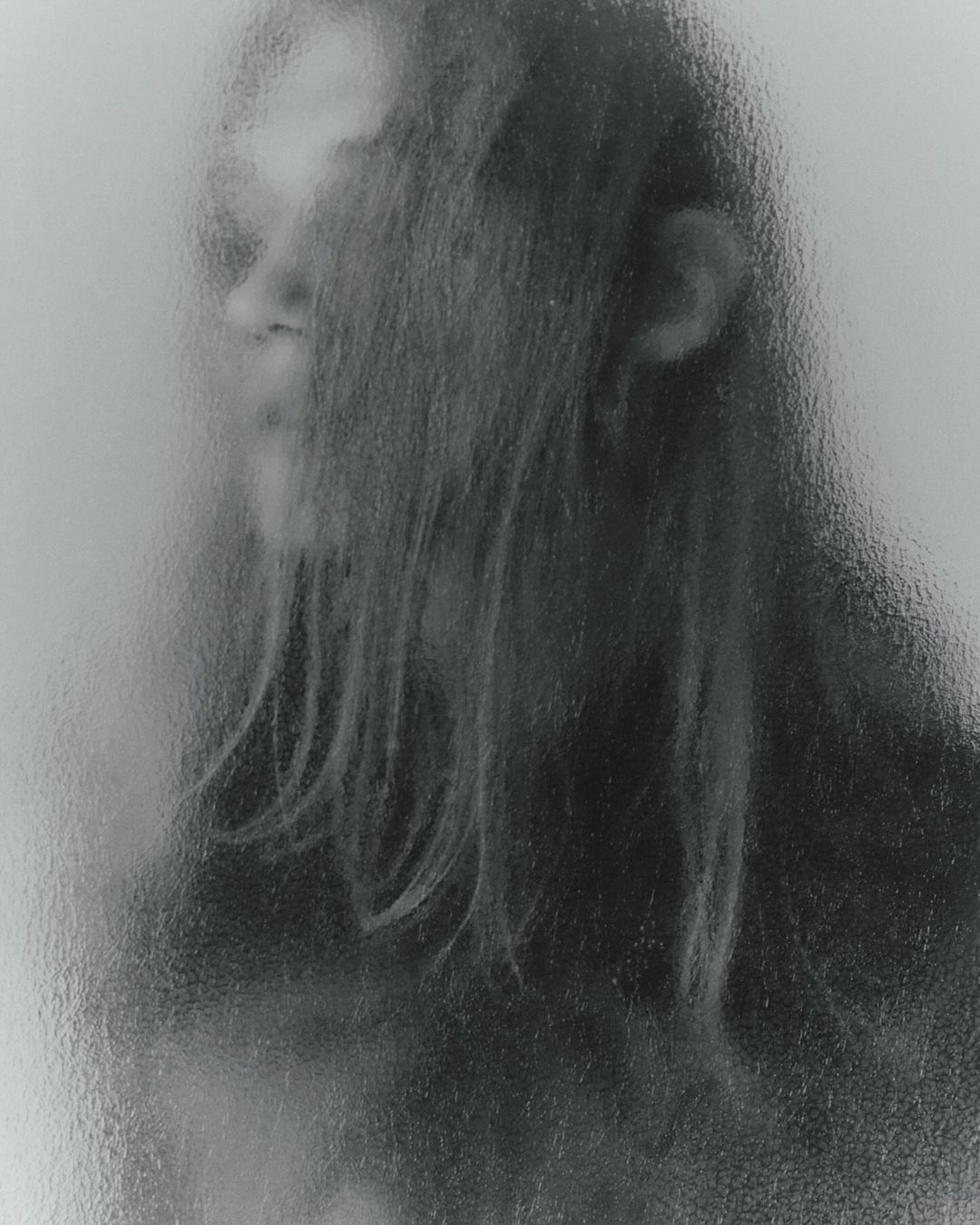 Monochromatic portrait of an individual with long hair, partially obscured by a textured glass or vapor effect, creating an ethereal mood.