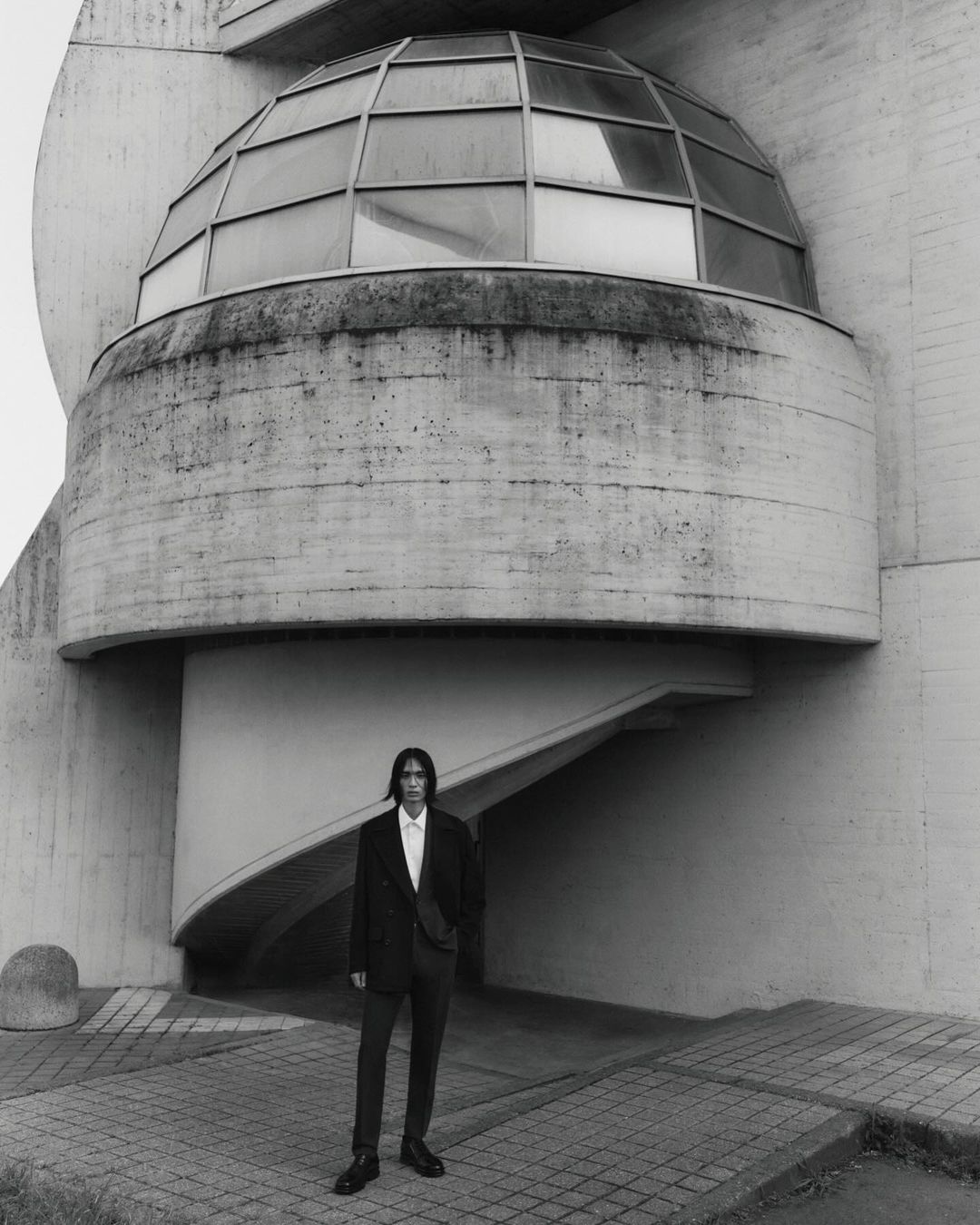 Monochrome shot of a male model in a suit posing in front of a modern architectural building featuring geometric shapes and a domed structure.